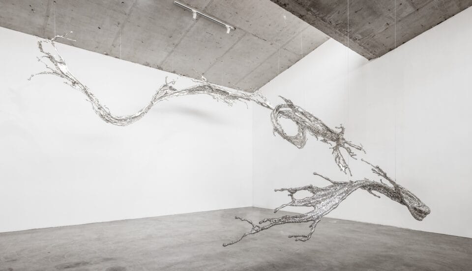 a chrome-colored abstract sculpture suspended from the ceiling in a gallery space that appears like a large splash of liquid frozen in time