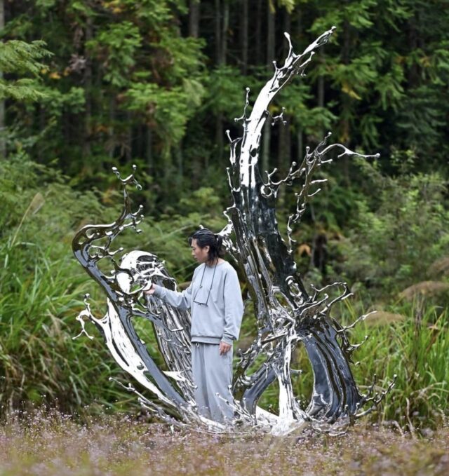 an Asian man stands at the base of a large, chrome-colored sculpture resembling a liquid splash frozen in time, installed in a field, with woods in the background