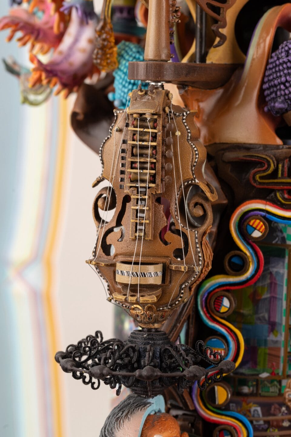 a close-up detail of part of an elaborately decorated sculpture with numerous sculpted, found, and painted elements, emphasizing an anthropomorphic stringed instrument with eyes and a mouth