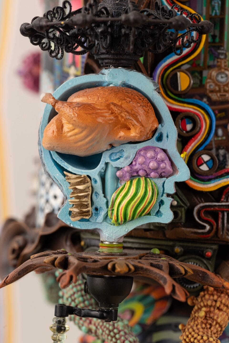 a close-up detail of part of an elaborately decorated sculpture with numerous sculpted, found, and painted elements, emphasizing a blue anatomical view of a human head with a chicken in the area where the brain should go and other colorful elements