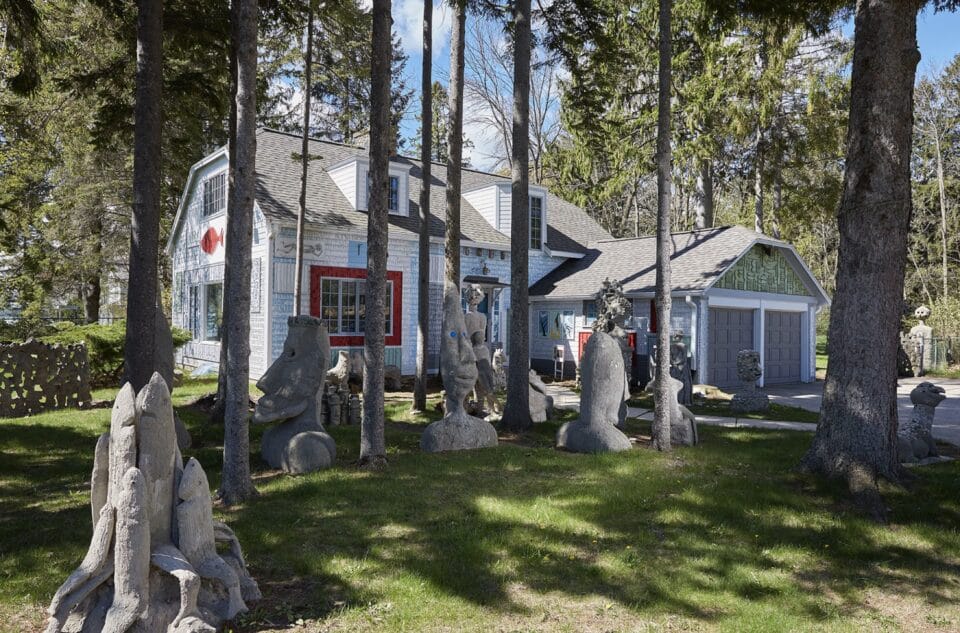 the exterior of artist Mary Nohl's house in southeastern Wisconsin, featuring a small white house among some pine trees with sculptures interspersed on the lawn