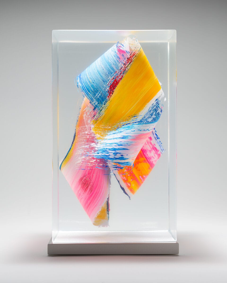 a sculpture made from cast clear resin in a cube form with three-dimensional multicolored brushstrokes inside, set against a gray background