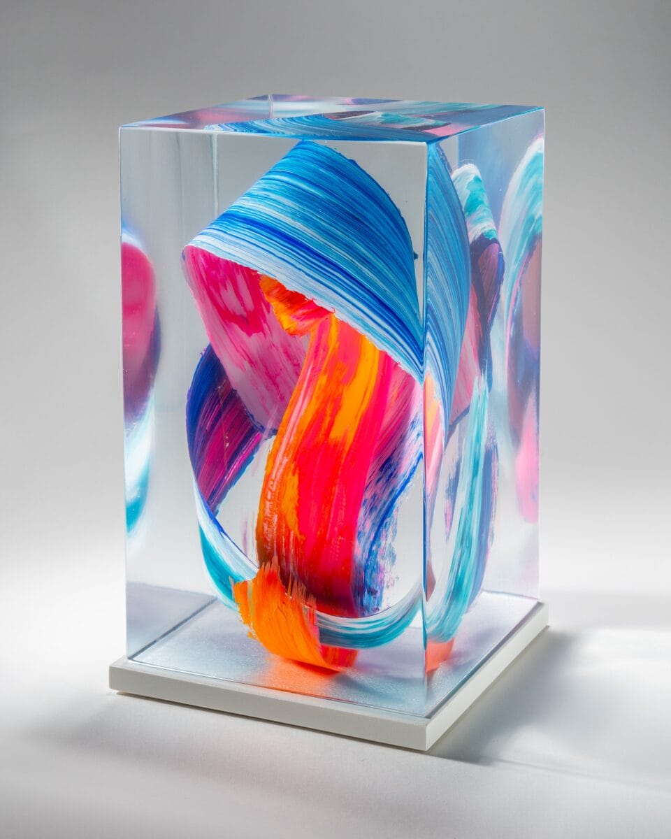 a three-quarter view of a sculpture made from cast clear resin in a cube form with three-dimensional multicolored brushstrokes inside, set against a gray background