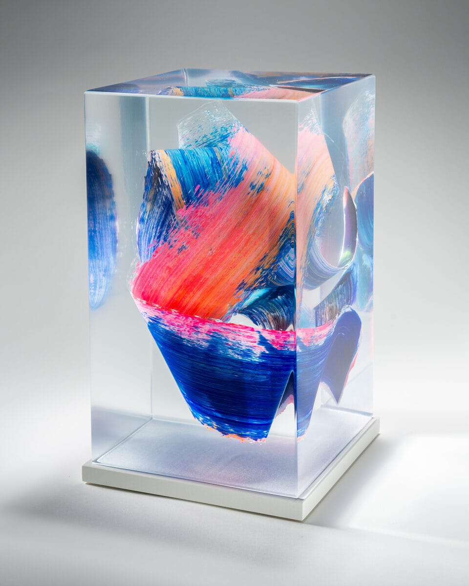 a three-quarter view of a sculpture made from cast clear resin in a cube form with three-dimensional multicolored brushstrokes inside, set against a gray background