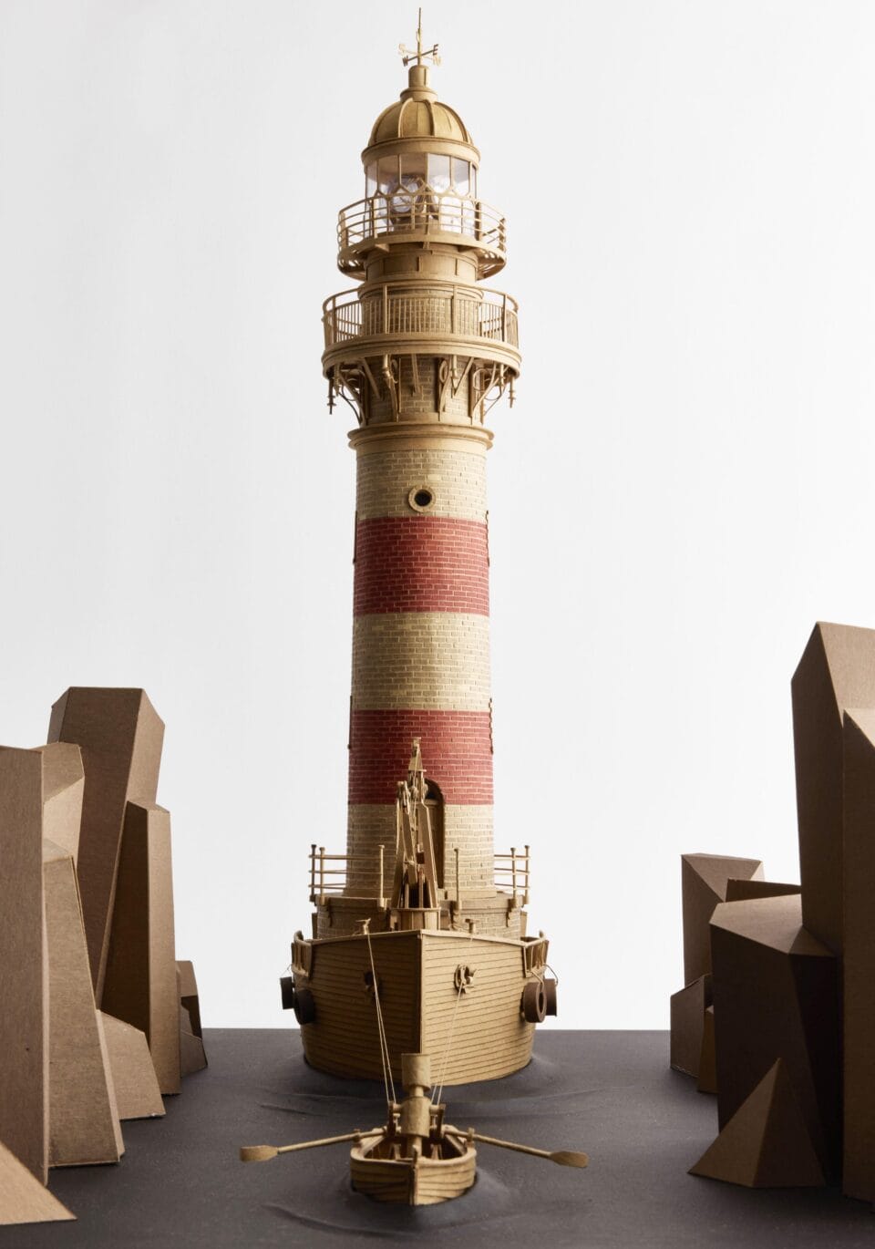 a cardboard sculpture of a lighthouse perched on top of a ship, being rowed through a rocky landscape by a small boat