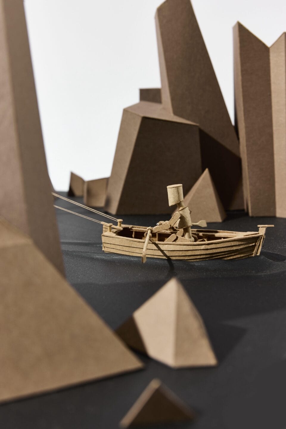 detail of a cardboard sculpture of a lighthouse perched on top of a ship, being rowed through a rocky landscape by a small boat