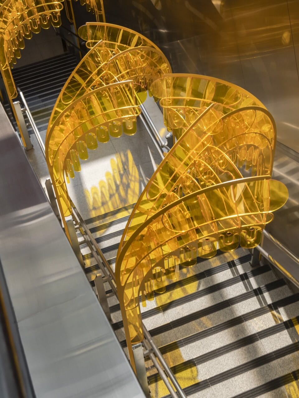 a yellow installation made of undulating pieces of arcylic, placed over a stairwell in a transit station, viewed from above