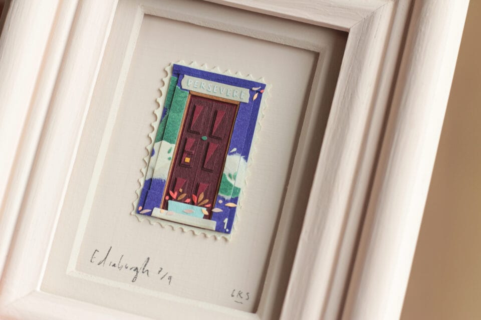 a hand holds up a small, vibrant collage in the shape of a stamp nestled inside a painted frame. it depicts a scene with a building and patterned designs