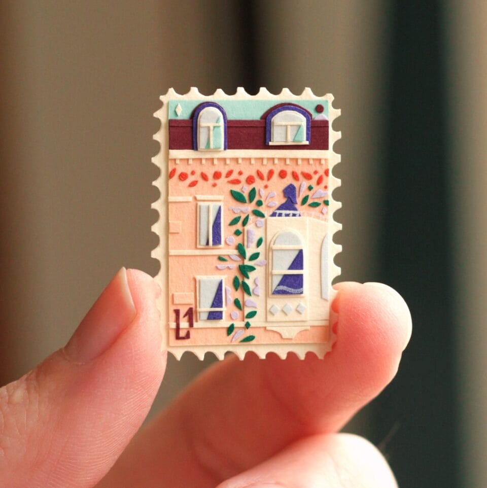 a hand holds up a small, vibrant collage in the shape of a stamp. it depicts a scene with a building and patterned designs