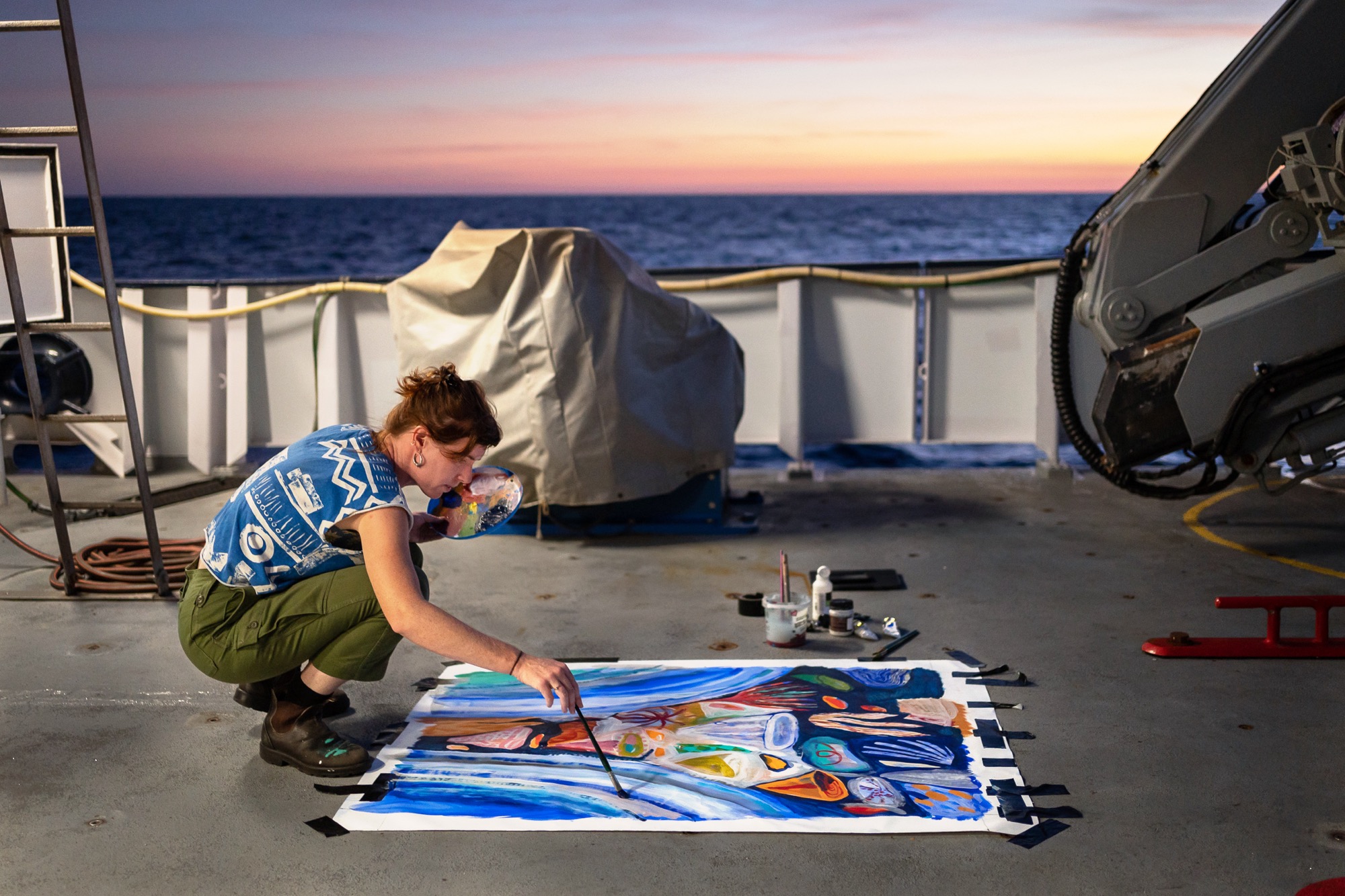 artist Ellie Hannon working on a painting on the aft deck of a research vessel at sunset