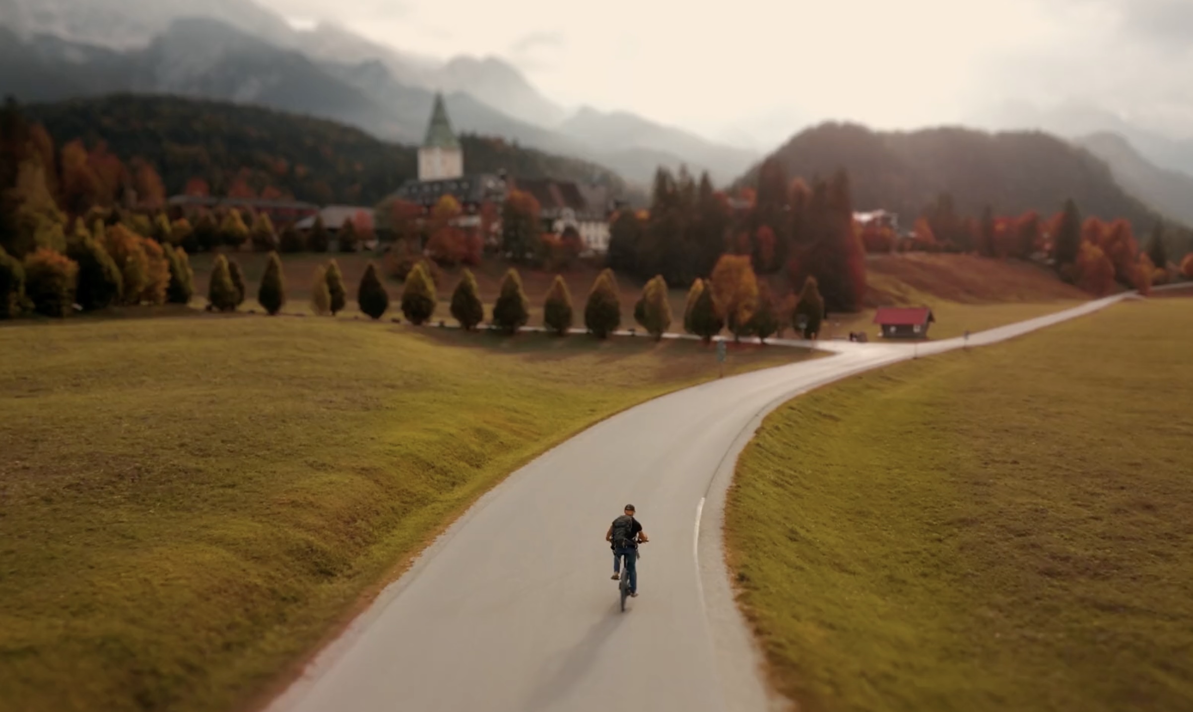 an aerial view of a cyclist on a road in an Alpine Valley with a historic hotel in the background