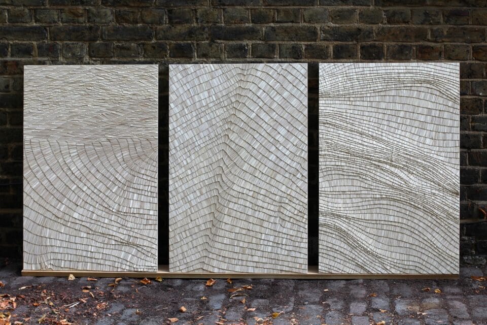 an abstract triptych artwork in a long, horizontal format, made from tiny, light gray, wooden shingles