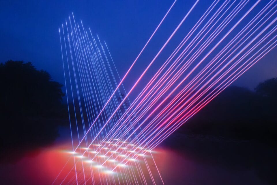 a landscape at dusk with a body of water that has geometric beams of light projected onto it in intersecting pink and blue lines, creating a grid of bright lights on the surface of the water