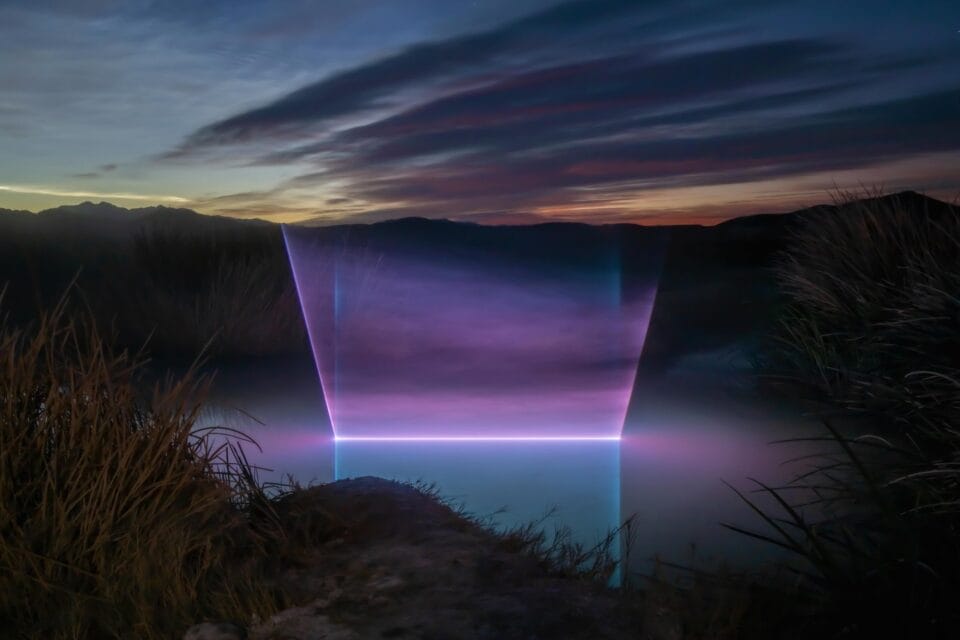 a landscape at dusk with a body of water that has a geometric beam of light projected onto it in a bright purple line