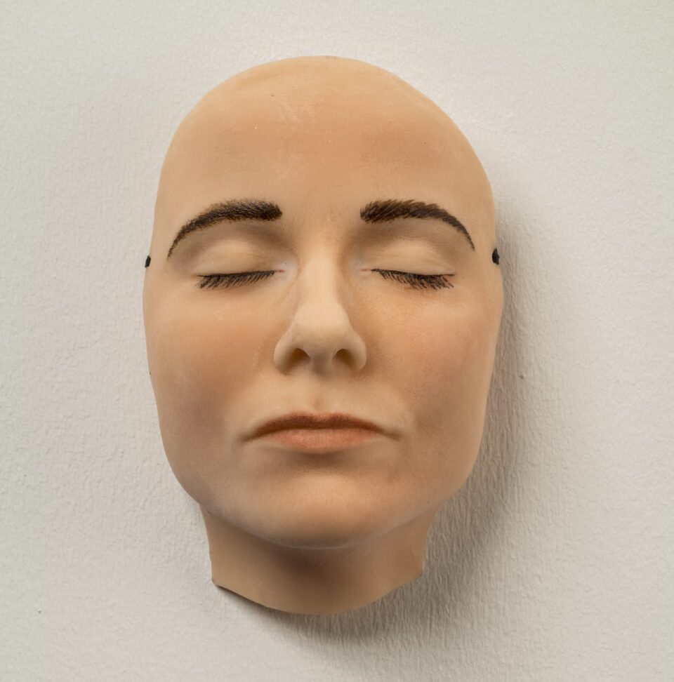 A highly realistic mask of a light-skinned face with dark brown eyebrows and eyelashes that appears to be sleeping—its eyes and mouth are closed and relaxed. The mask hangs on a white wall.