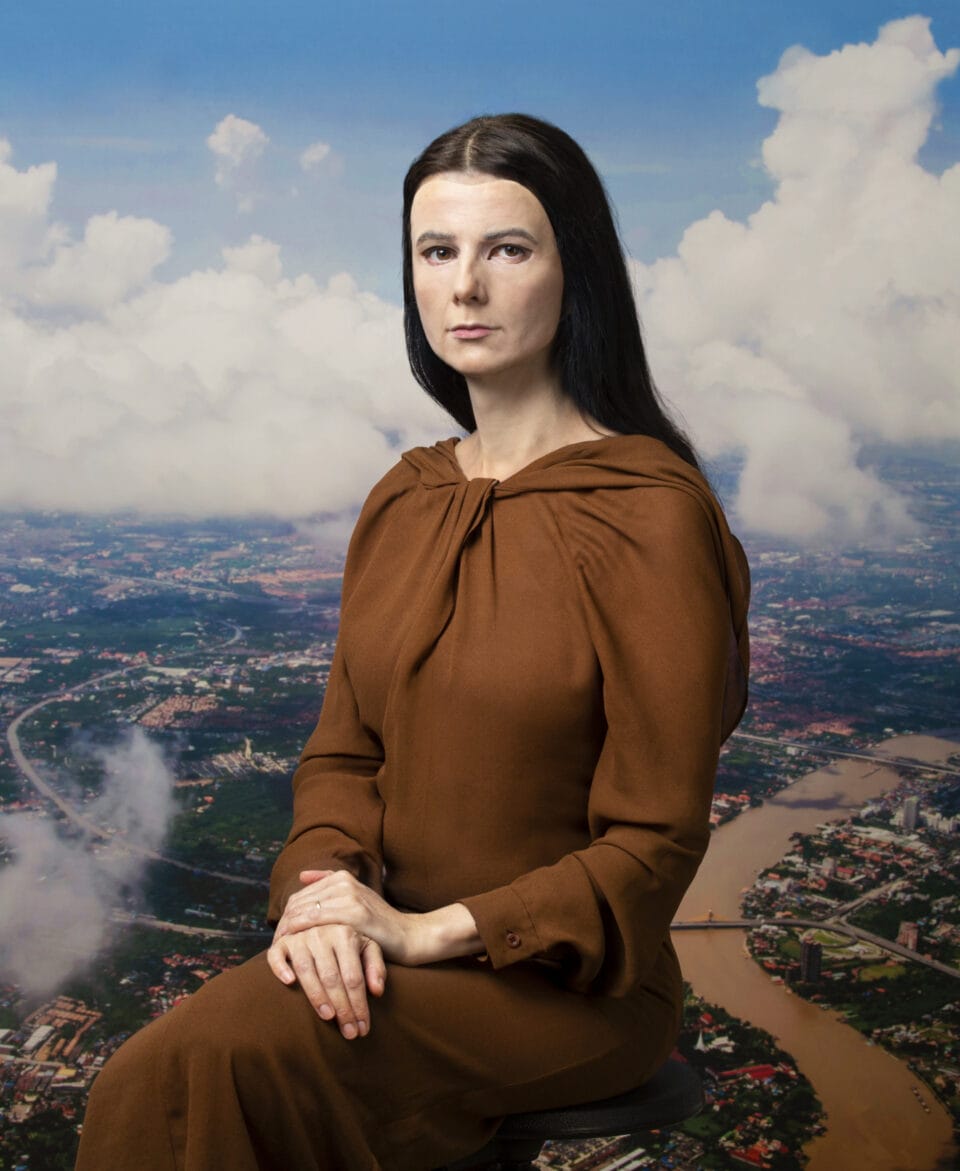 A light-skinned woman wears a realistic mask of another woman with the same skin tone. She sits posed like the Mona Lisa. She has long dark hair and wears a brown dress. The backdrop behind her shows a cityscape with rivers and clouds.